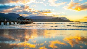 Un muelle que se adentra en el mar, con una estructura techada al final, rodeado de montañas y un cielo al atardecer.