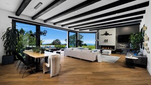 Un salon moderne doté d’un plancher en bois, d’un canapé blanc, d’une table à manger avec des chaises et de grandes fenêtres offrant une vue sur l’extérieur.