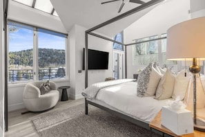 A modern bedroom with a large bed, a chair, a TV, and a view of a forest.