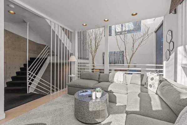 A modern living room with a staircase, a grey sofa set, a round coffee table, and a view of bare trees through large windows.