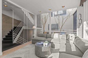 Uma sala de estar moderna com escada, um sofá cinza, uma mesa de centro redonda e vista para árvores secas através de grandes janelas.