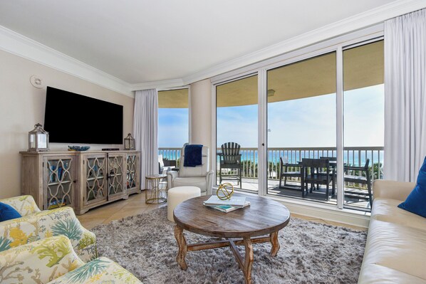 A spacious living room with a large TV, a wooden coffee table, and a sofa. There is a view of the ocean through large sliding doors.