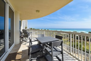 A balcony with a table and chairs, overlooking a beach with a clear view of the ocean and a distant building.