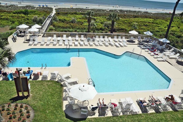 A large pool with lounge chairs and umbrellas, surrounded by greenery and a beach view.