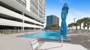 A swimming pool with lounge chairs and an umbrella, surrounded by a white building with balconies. - Regency Isle 507 by Liquid Life (Orange Beach)