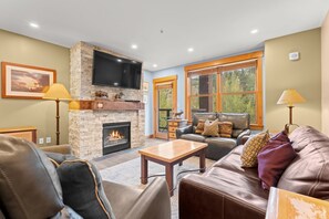 A living room with a stone fireplace, leather furniture, a flat-screen TV, and large windows with a view of trees.