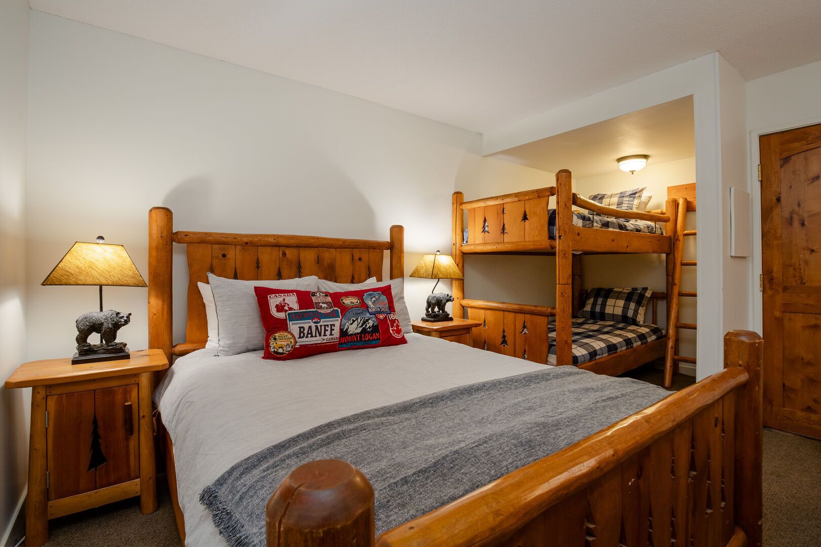 A room with a wooden bunk bed, a single bed with a Canada-themed pillow, two bedside tables with lamps, and a wardrobe.