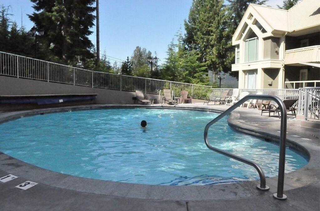 A swimming pool with a metal ladder, surrounded by lounge chairs and a building with a balcony.