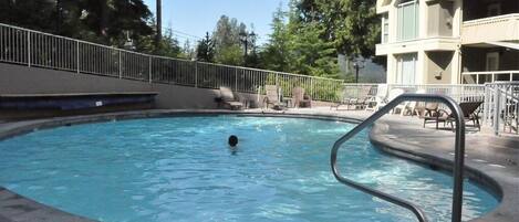 A swimming pool with a metal ladder, surrounded by lounge chairs and a building with a balcony.