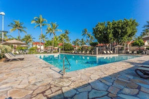 A swimming pool with stone flooring, lounge chairs, umbrellas, and palm trees.