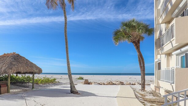 Vista de la playa con palmeras, una cabaña de techo de paja y un edificio con balcones.