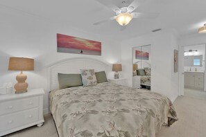 Une chambre à coucher comprenant un lit, des tables de chevet, un ventilateur de plafond, un tableau, un miroir, ainsi qu’une salle de bain avec un lavabo et des placards.