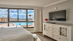 A hotel room with a large window, a flat-screen TV, a bed, a dresser, and a view of a pool and buildings. - WALK OUT TO POOL FROM FURNISHED PATIO!  Beachside Condo, Just Steps to Beach! (Miramar Beach)