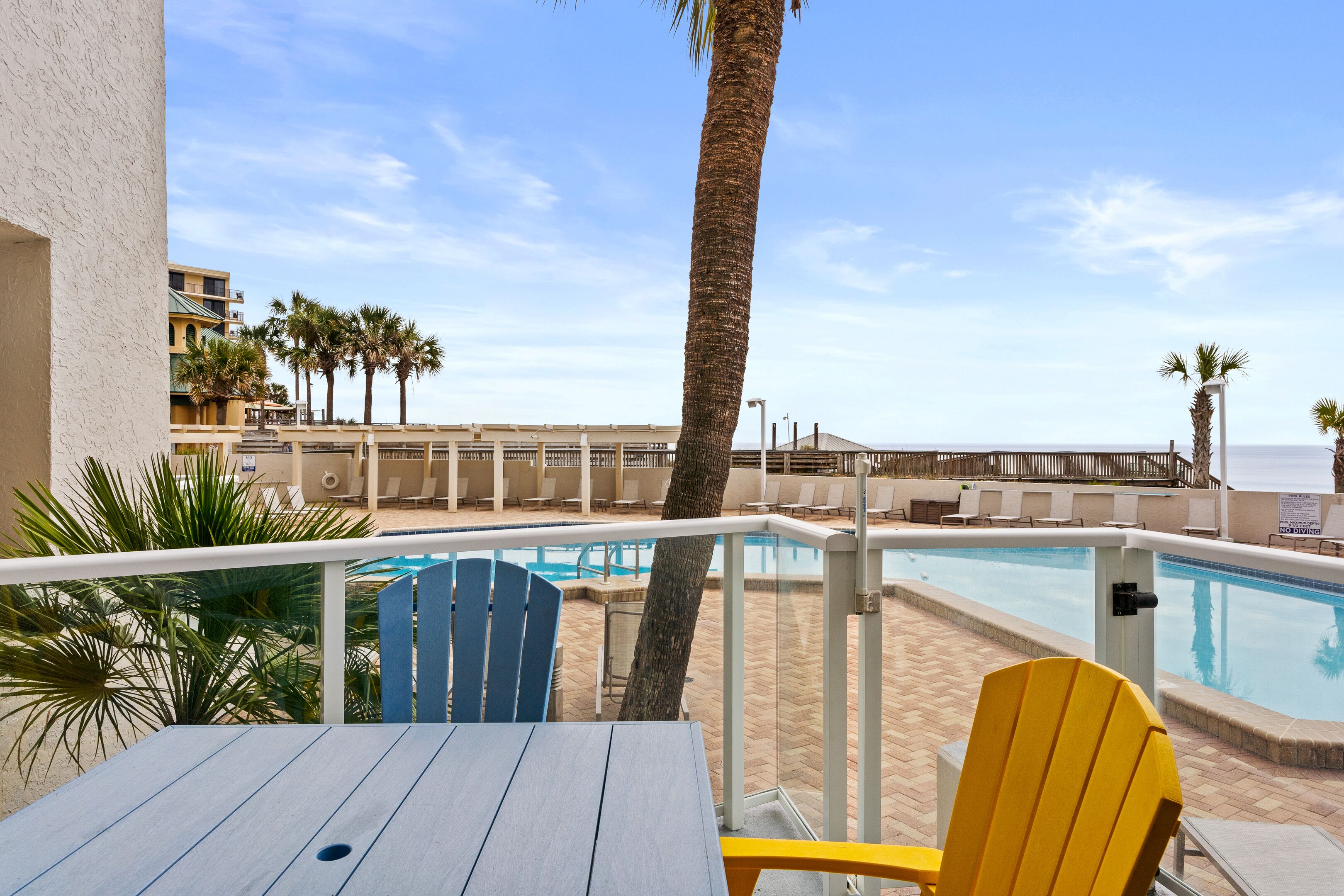A poolside area with a table and chairs, a palm tree, and a view of the beach.