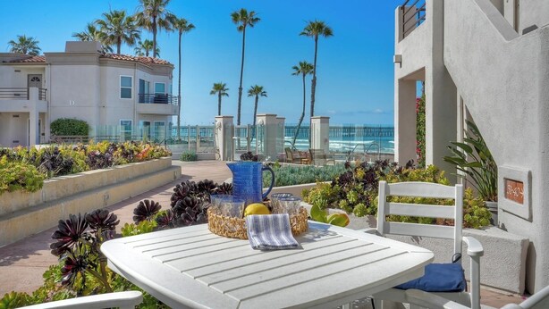 A patio with a table and chairs, a railing, and a view of the ocean and buildings. - Premium Beachfront Stay Ground Floor, 2 Parking Spots,  Community Jacuzzi EV Charging (Oceanside)