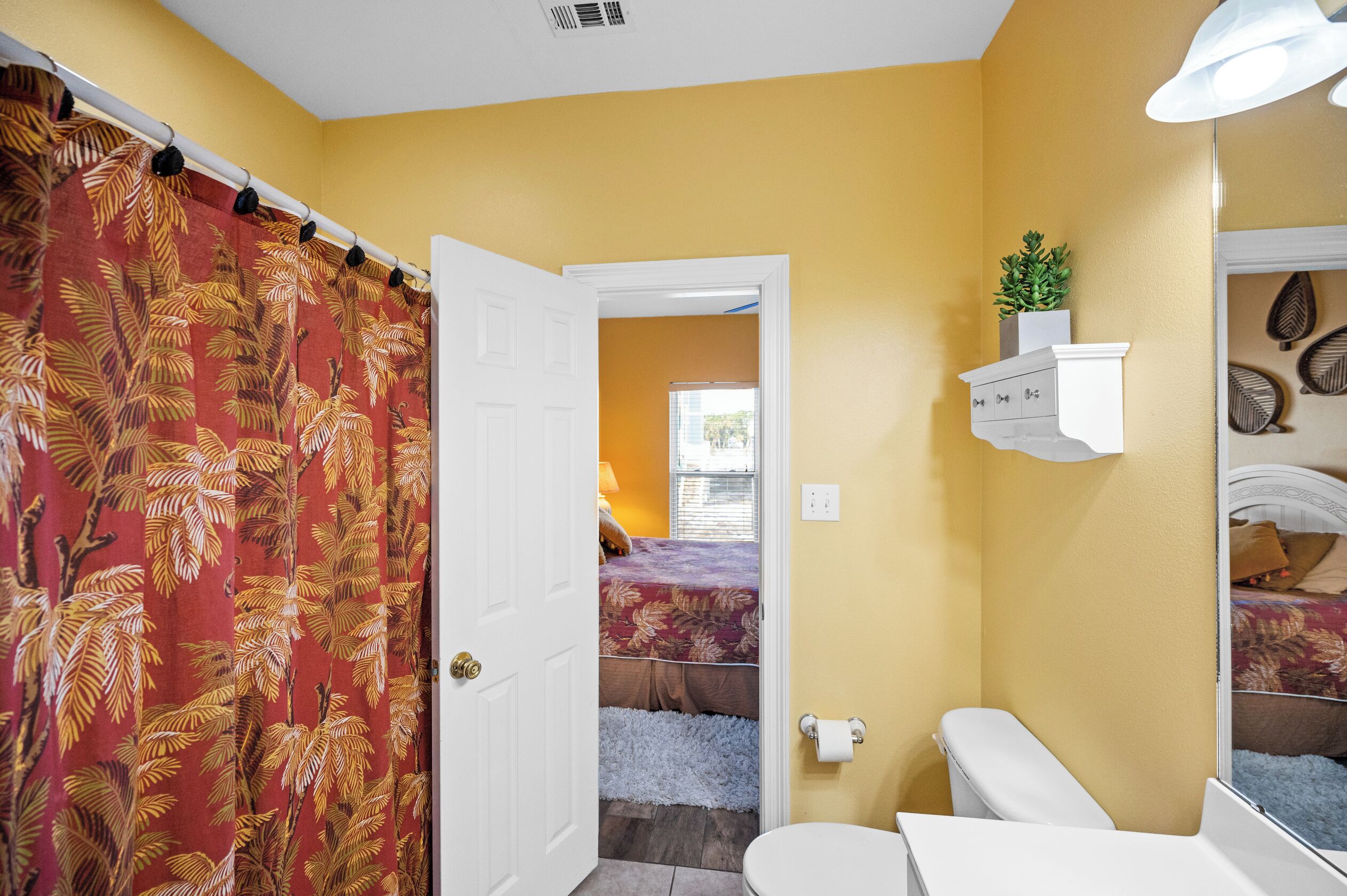 Une salle de bain avec un mur jaune, des toilettes blanches, un rideau de douche orné de motifs de feuilles et une petite étagère avec une plante.