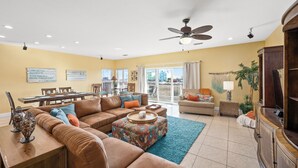 A spacious living room with a brown leather sofa, a coffee table, and a dining area with a table and chairs. There is a ceiling fan, a TV, and a large window with curtains.