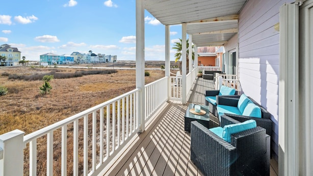 A covered porch with outdoor seating, overlooking a beach and buildings.
