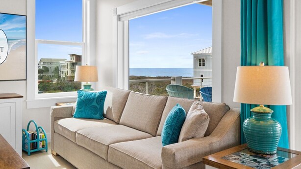Una sala de estar con un sofá color crema, cojines verde mar, dos lámparas y vistas al océano y a las casas a través de amplios ventanales.