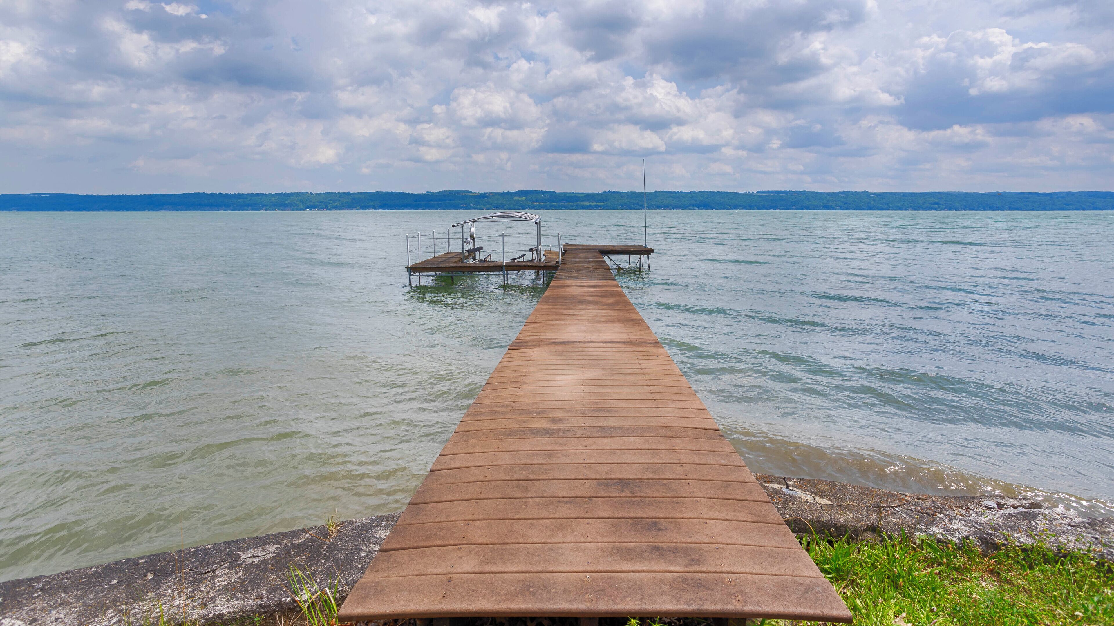 Une jetée en bois s’étendant dans un lac paisible, avec une rive lointaine et un ciel nuageux.