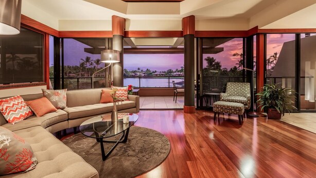 Een moderne woonkamer met een gebogen bank, een glazen salontafel en uitzicht op een zonsondergang over het water.