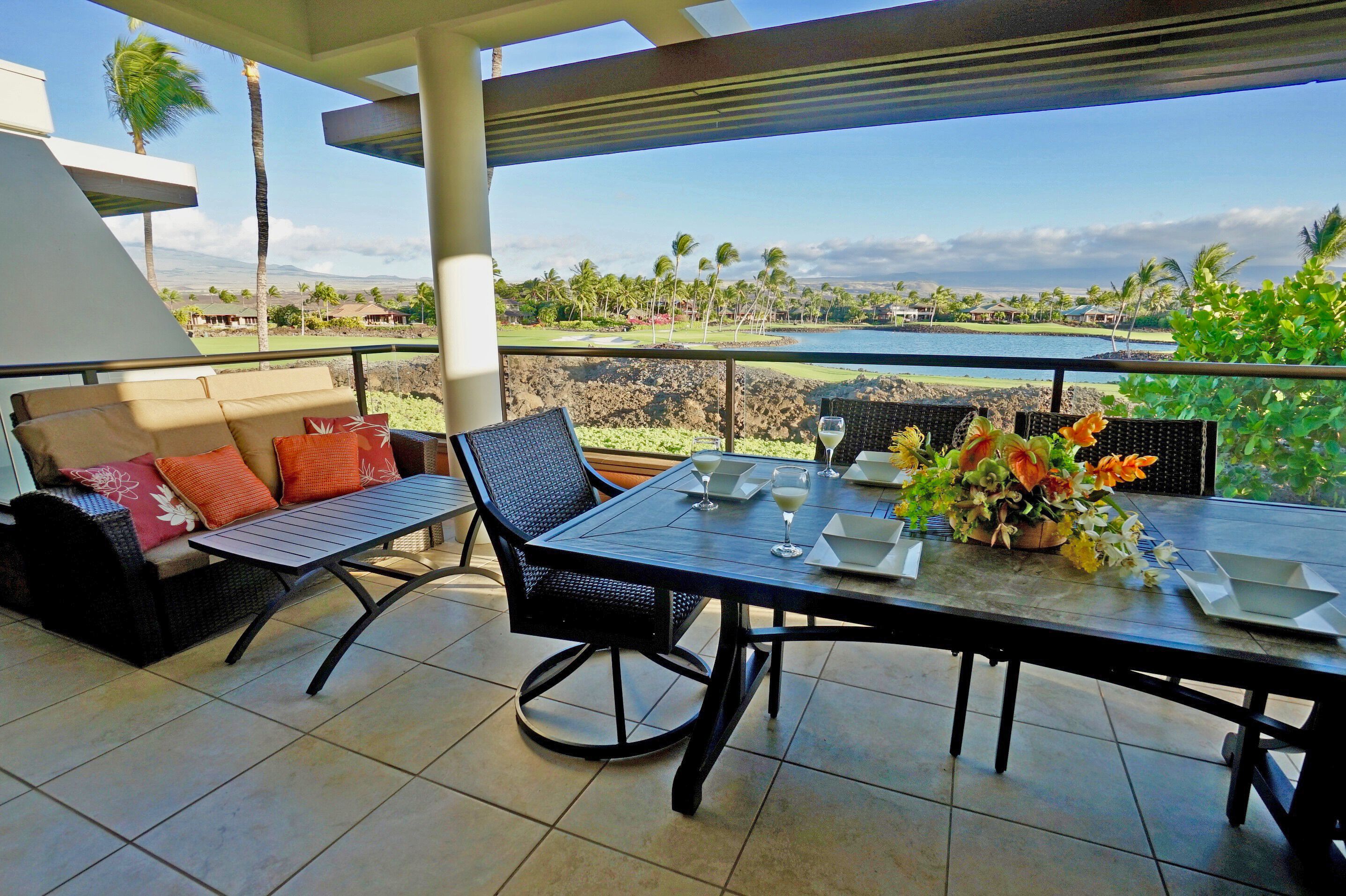 A terrace with a dining table set for two, a sofa with cushions, and a view of the ocean and palm trees.