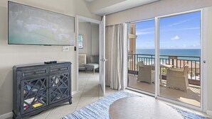 Quarto de hotel com uma televisão grande, um console de madeira escura, uma varanda com mobiliário de vime e vista para o oceano.