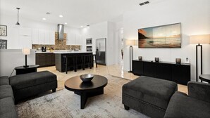 A modern living room with a large black sofa, a round coffee table, and a kitchen area in the background. - EV_116 - 6BR / 4.5BA Highlands Reserve Villa near Disney, Private Pool & Spa (Davenport)