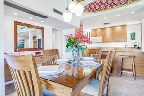 A dining area with a wooden table, chairs, a vase with flowers, and a kitchen in the background.