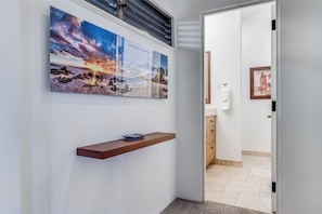 A hallway with a wooden shelf, a wall-mounted artwork depicting a sunset, and a door leading to a bathroom.