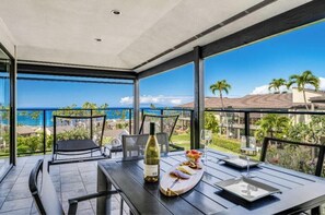 A balcony with a table set for dining, a bottle of wine, and a view of the ocean and palm trees.