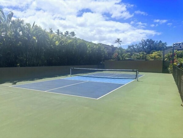 An outdoor tennis court with a net, surrounded by greenery and a clear sky.