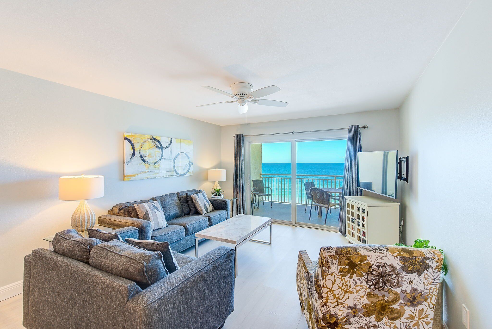 A modern living room with a sofa, armchair, and coffee table, a ceiling fan, and a view of the ocean through large sliding doors.