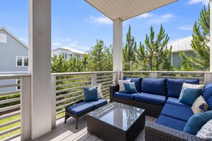 A covered outdoor seating area with a sofa and a glass-top coffee table, surrounded by white railings and greenery.