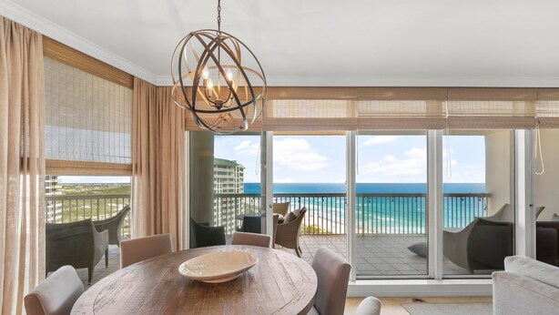 A dining area with a round wooden table, beige chairs, and a large window with a view of the ocean and buildings. - WELL APPOINTED & ELEGANT CONDO! GULF FRONT WITH SWEEPING VIEW (Destin)