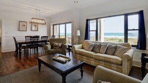 A spacious living room with a large window overlooking the beach, a dining area with wooden furniture, and a ceiling fan. - DR 2211–Simply elegant oceanfront condo with expansive views, pool and tennis (Wrightsville Beach)