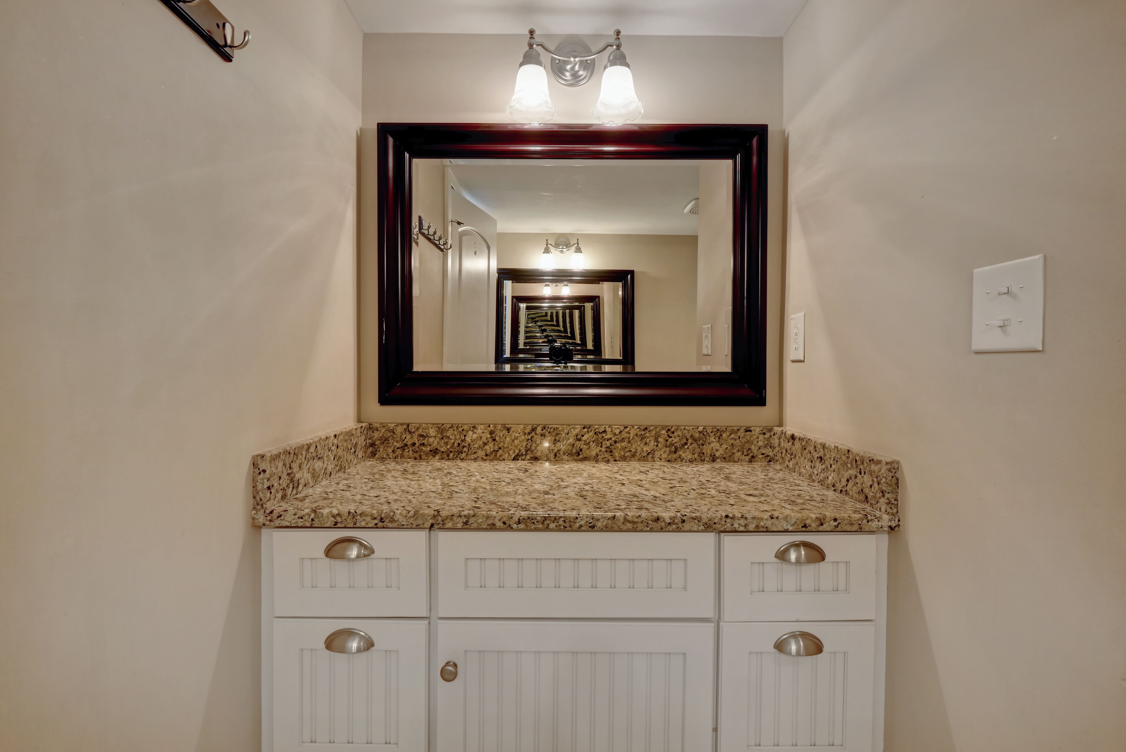 Un baño con encimera de granito, muebles blancos, un espejo grande y un pasillo visible en el reflejo.