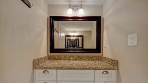 A bathroom with a granite countertop, white cabinetry, a large mirror, and a hallway visible in the mirror.