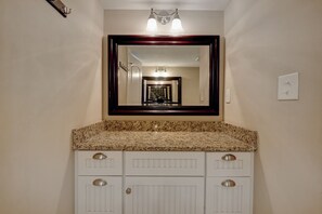 A bathroom with a granite countertop, white cabinetry, a large mirror, and a hallway visible in the mirror.