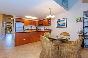 A kitchen with wooden cabinets, a refrigerator, a microwave, and a dining area with wicker chairs and a glass table.