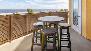 A balcony with a round table and two stools, overlooking a beach with palm trees and the ocean. - Sandpiper 203 | Oceanfront with Great Views! (Tybee Island)