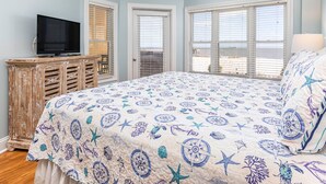 A bedroom with a large bed, a television, and a view of the beach through the windows. - Sandpiper 203 | Oceanfront with Great Views! (Tybee Island)