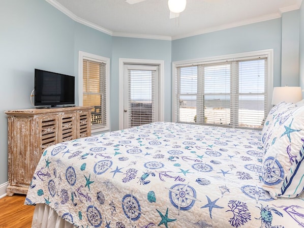 A bedroom with a large bed, a television, a wooden dresser, and a view of the beach through the windows.