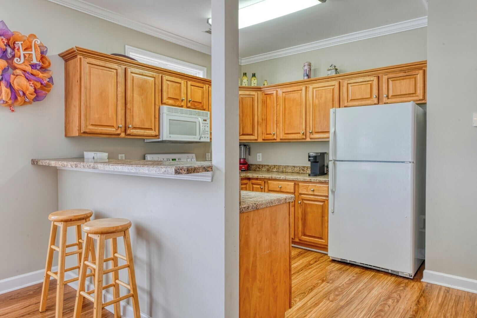 A kitchen with wooden cabinets, a white refrigerator, a microwave, and a coffee maker. There are two wooden stools and a decorative wall hanging.