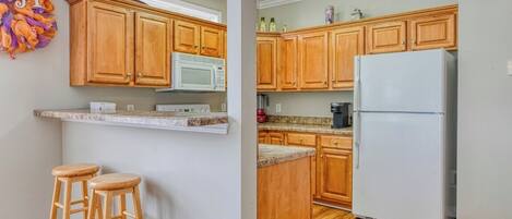 A kitchen with wooden cabinets, a white refrigerator, a microwave, and a coffee maker. There are two wooden stools and a decorative wall hanging.