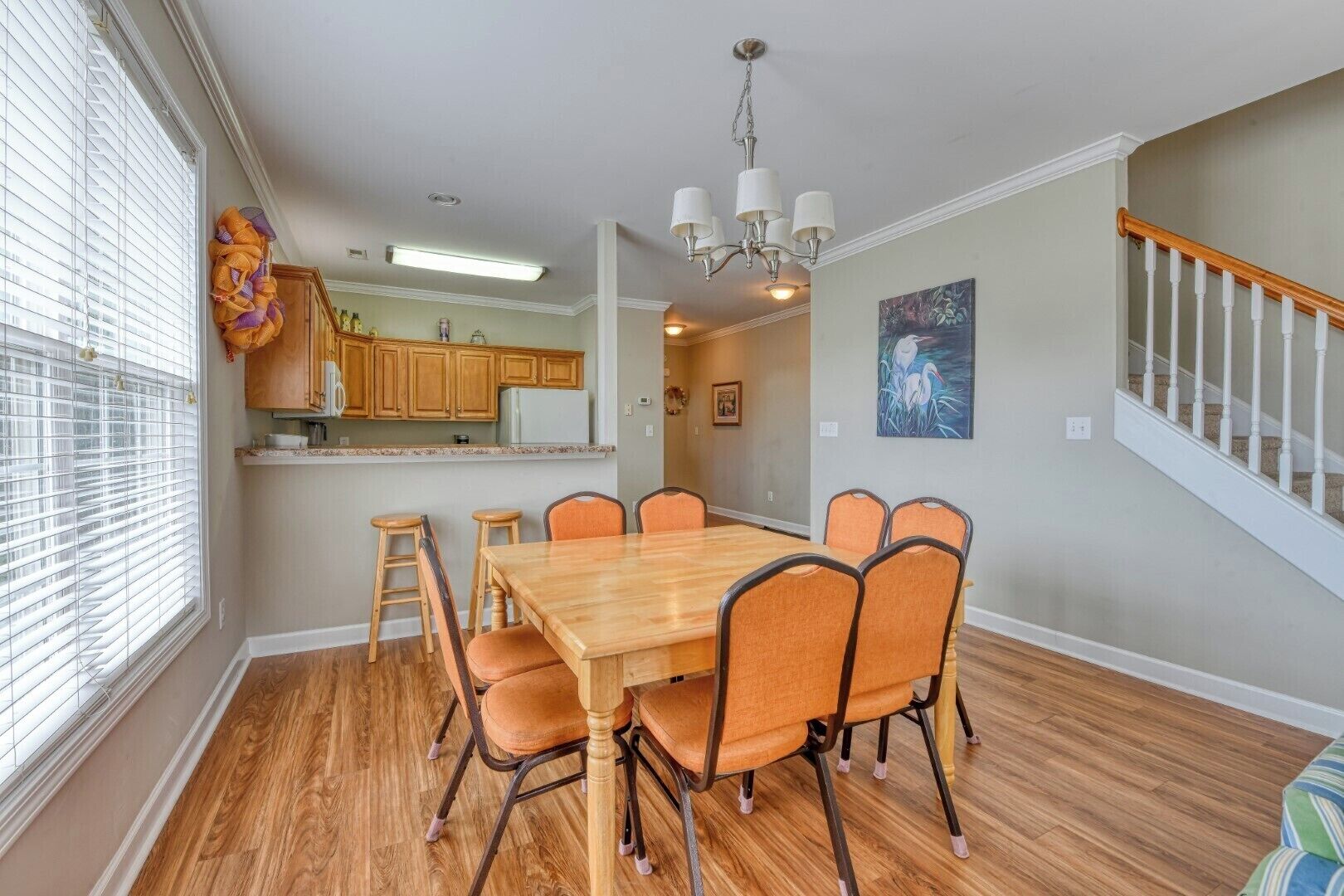 A dining room with a wooden table and chairs, a chandelier, and a view of the kitchen and staircase.