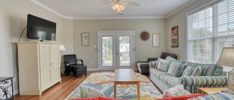 A living room with a sofa set, a television on a cabinet, a ceiling fan, and a door leading outside.
