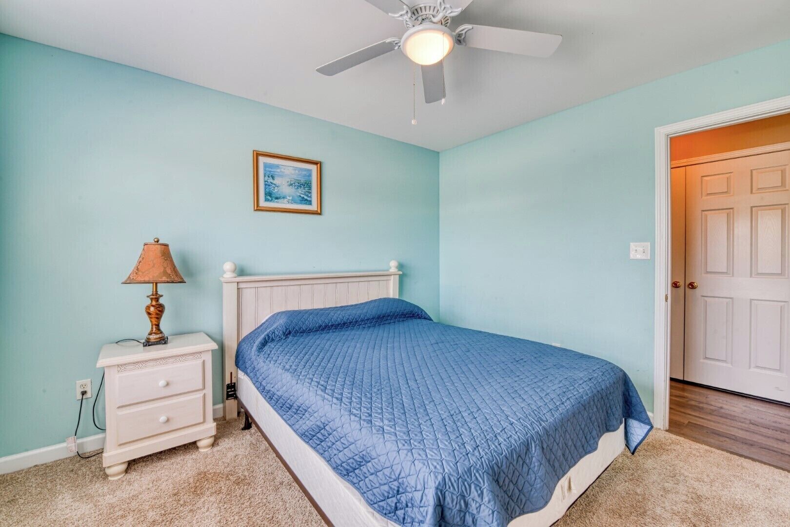A bedroom with a bed, a nightstand, a lamp, a ceiling fan, and a door.