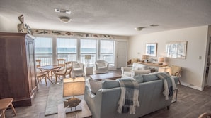 A spacious living room with a large grey sofa, a dining table with chairs, and a balcony with large windows. - Greenwave - Gulf Front, Old Florida Style on Dune Allen Beach (Santa Rosa Beach)
