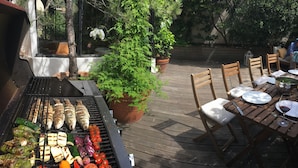 Ein Grill im Freien mit verschiedenen Fleischsorten und Gemüse, die zubereitet werden. Eine Holzterrasse mit Stühlen und einem Tisch ist für das Essen gedeckt.